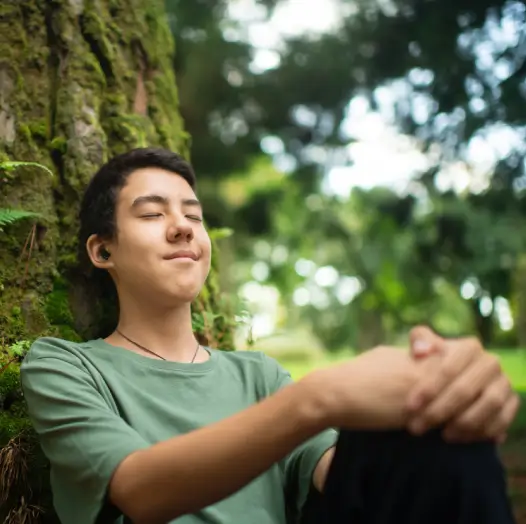 A young man sitting against a tree and listening to music while meditating, an approach to holistic mental health treatment